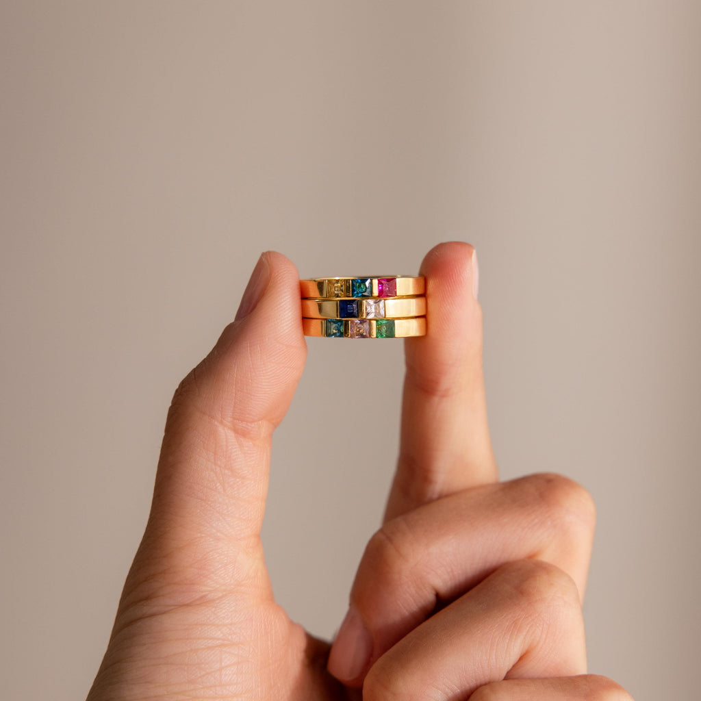 A hand displays three stacked Multiple Birthstone Bands with colorful gemstones, highlighting a personalized jewelry style—ideal as a unique birthstone ring—against a plain background.