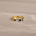 The Multiple Birthstone Band features five colorful square gemstones set in a row on a soft beige surface.
