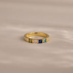 The Multiple Birthstone Band features five colorful square gemstones set in a row on a soft beige surface.