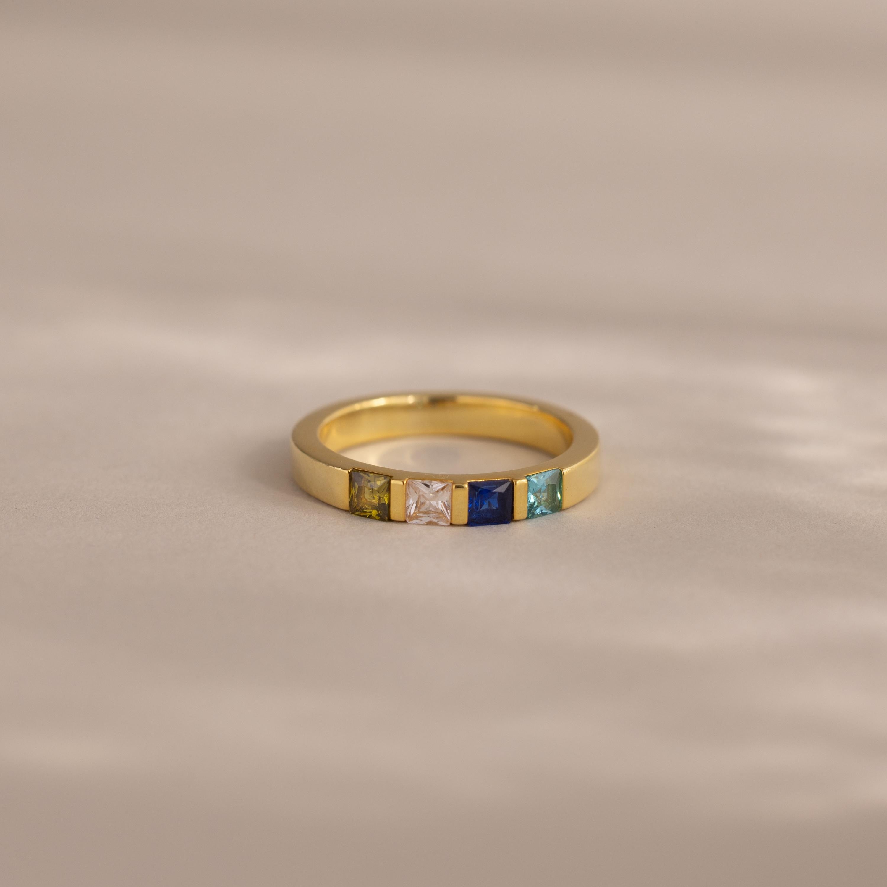 The Multiple Birthstone Band features five colorful square gemstones set in a row on a soft beige surface.