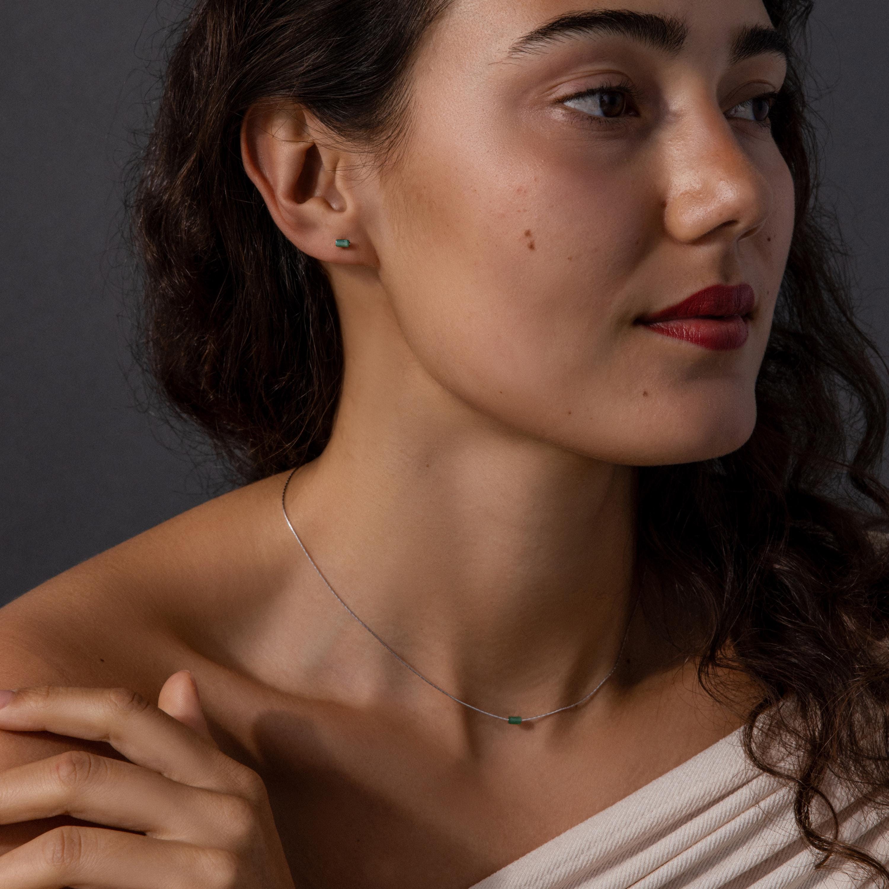 Woman with wavy hair wears the Jade Bar Jewelry Set in Sterling Silver, featuring a silver necklace and small green stud earrings, as she looks to the side.