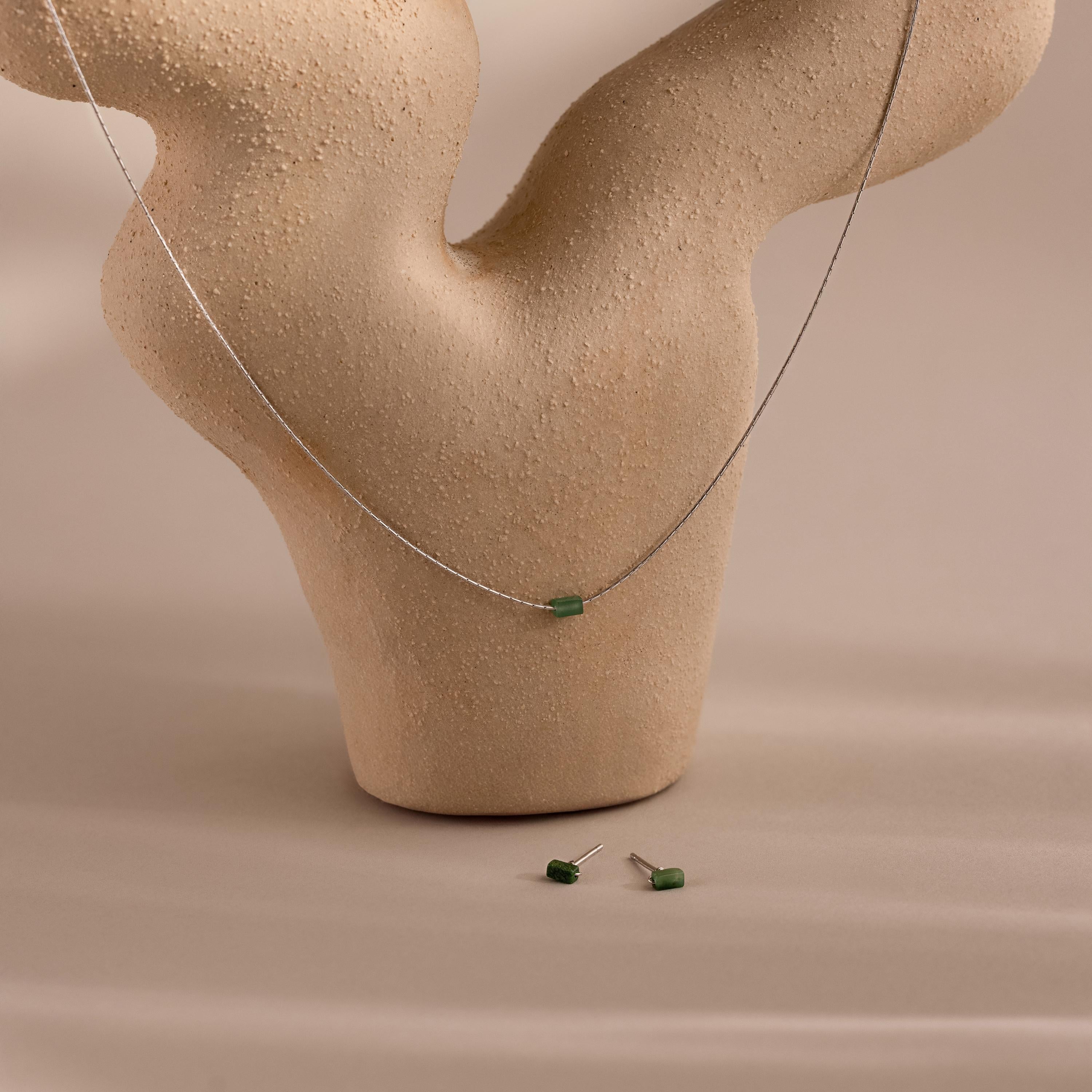 The Jade Bar Jewelry Set in Sterling Silver, featuring a bar necklace and stud earrings with small green jade stones, is displayed on a beige textured abstract stand.