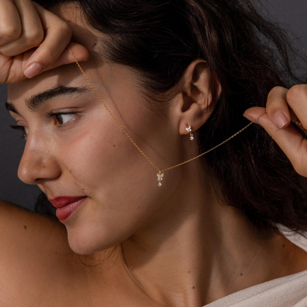 A woman holds the Diamond Butterfly Drop Jewelry Set necklace near her face, wearing matching diamond butterfly huggie earrings, and looks to the side.