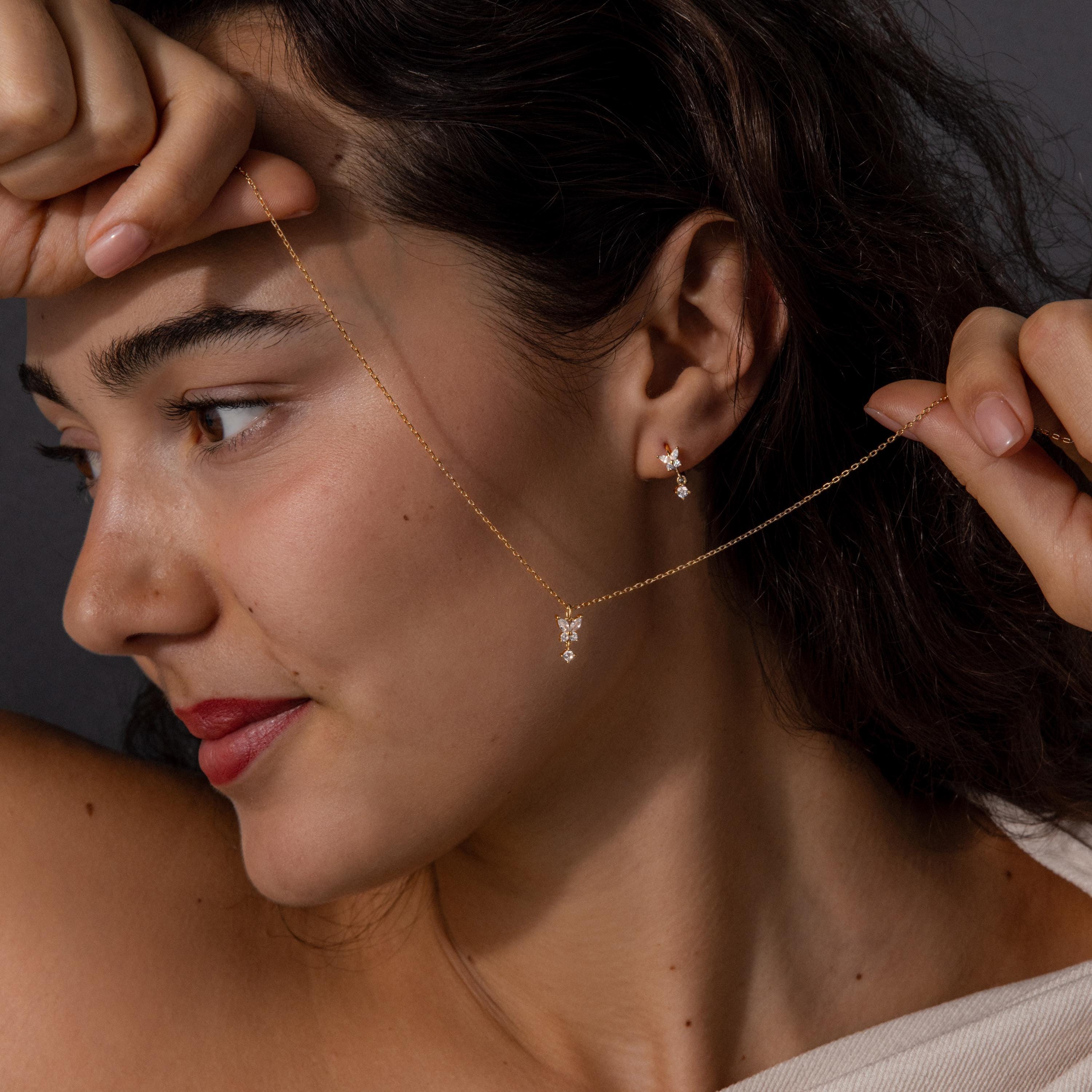 A woman holds the Diamond Butterfly Drop Jewelry Set necklace near her face, wearing matching diamond butterfly huggie earrings, and looks to the side.