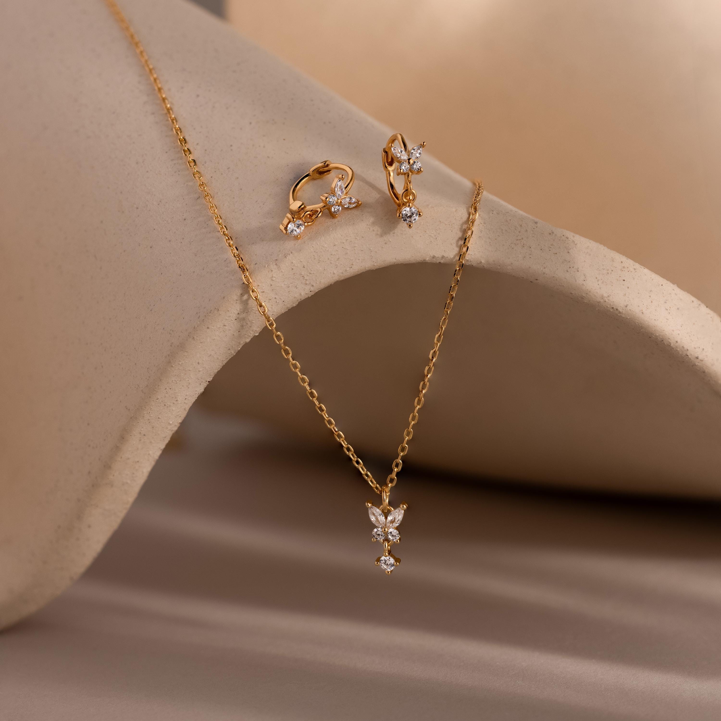 The Diamond Butterfly Drop Jewelry Set, featuring sparkling huggie earrings and a butterfly drop necklace, is elegantly displayed on a neutral curved surface.