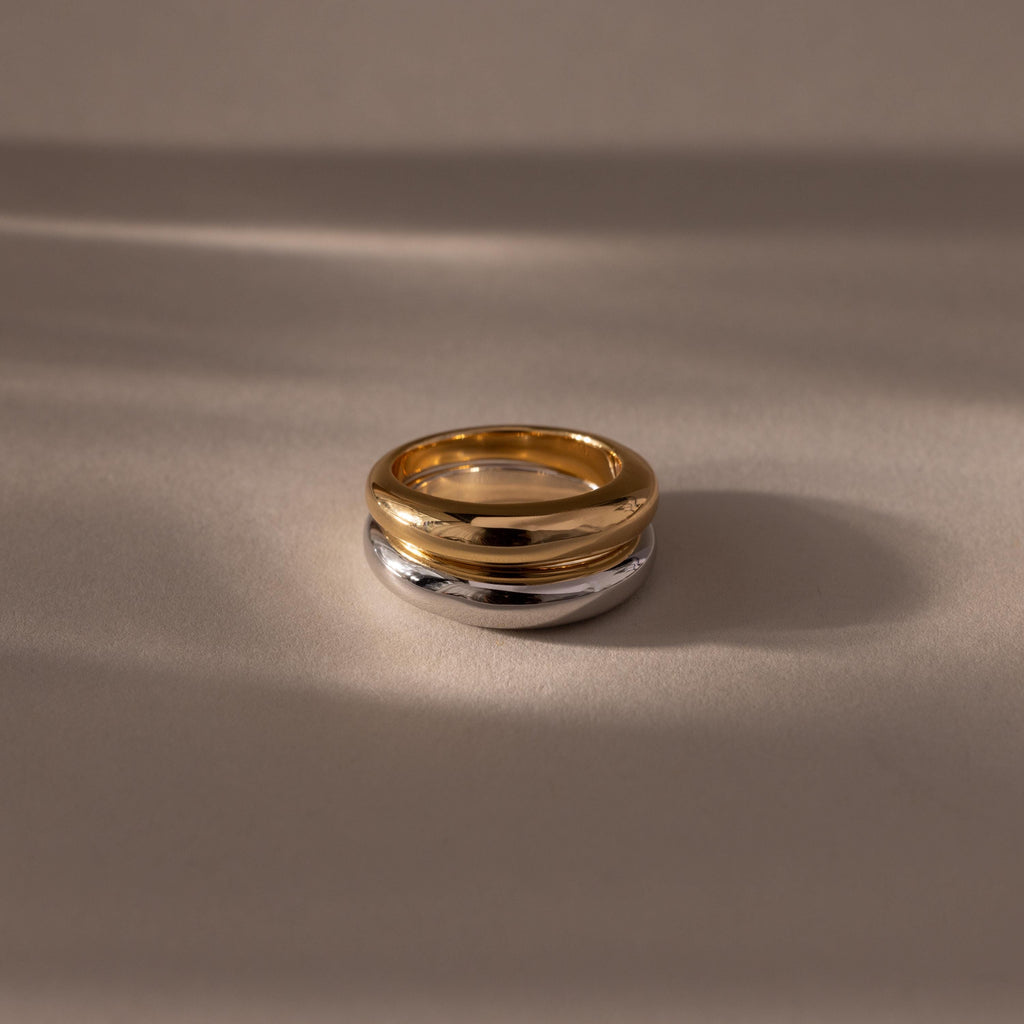 The Mixed Metal Dome Ring Set features a gold and silver ring intertwined, resting on a smooth beige surface with soft shadows—a versatile and modern classic jewelry piece.