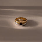 The Mixed Metal Dome Ring Set features a gold and silver ring intertwined, resting on a smooth beige surface with soft shadows—a versatile and modern classic jewelry piece.