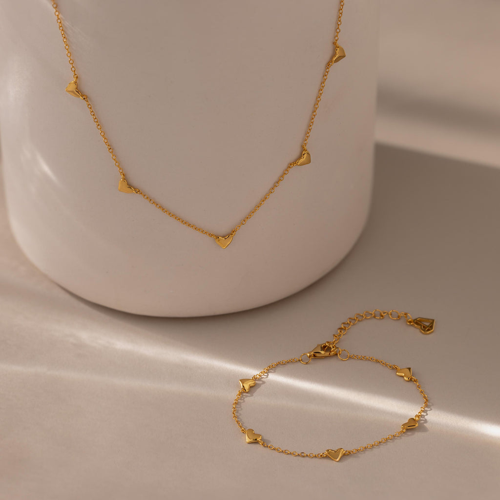 The Heart Station Necklace & Bracelet Set in 18K Gold features delicate heart charms, beautifully displayed on a neutral ceramic surface.