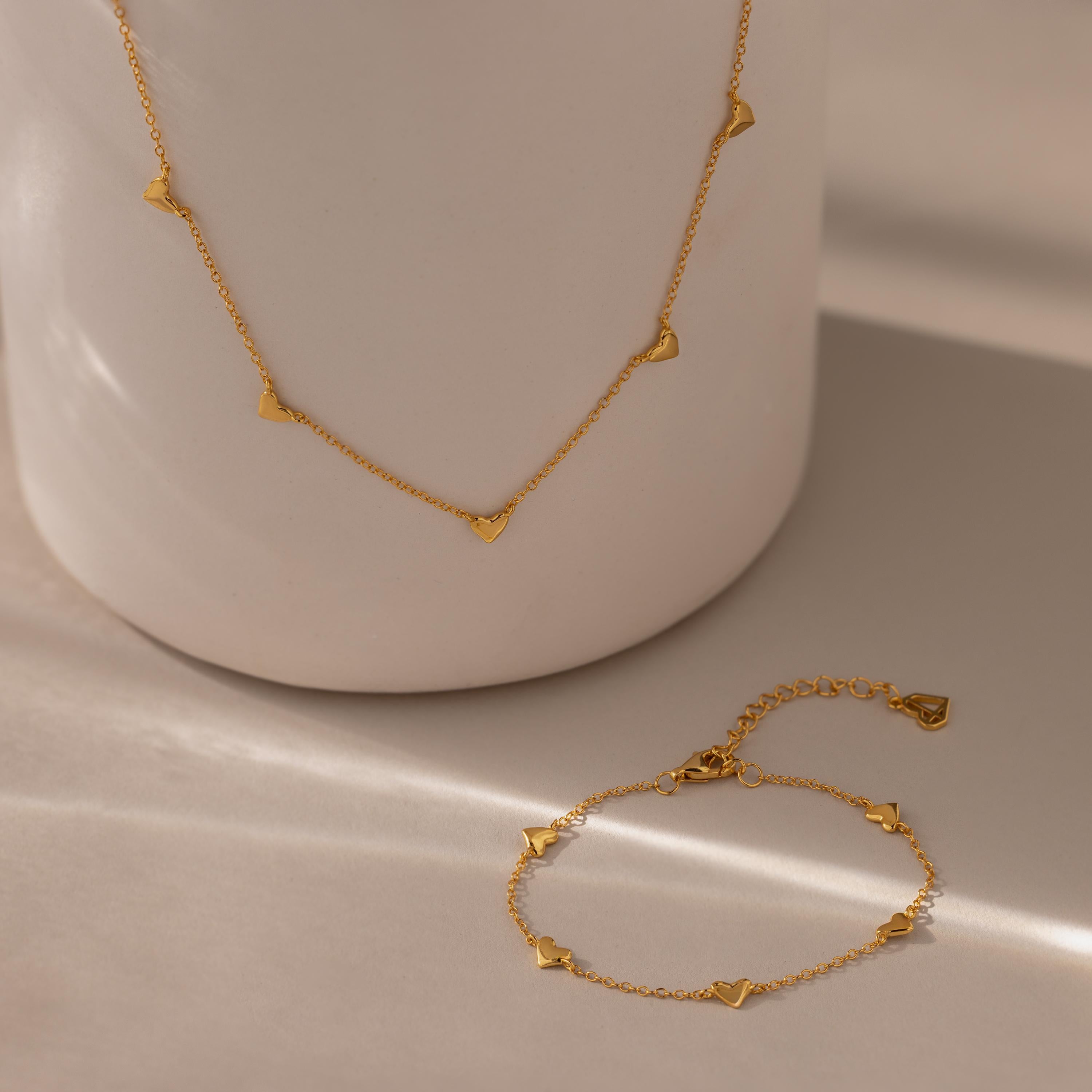 The Heart Station Necklace & Bracelet Set in 18K Gold features delicate heart charms, beautifully displayed on a neutral ceramic surface.