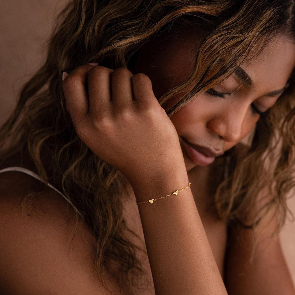 Woman with wavy hair wearing the Heart Station Necklace & Bracelet Set in 18K Gold, featuring delicate heart charms, resting her head on her hand.
