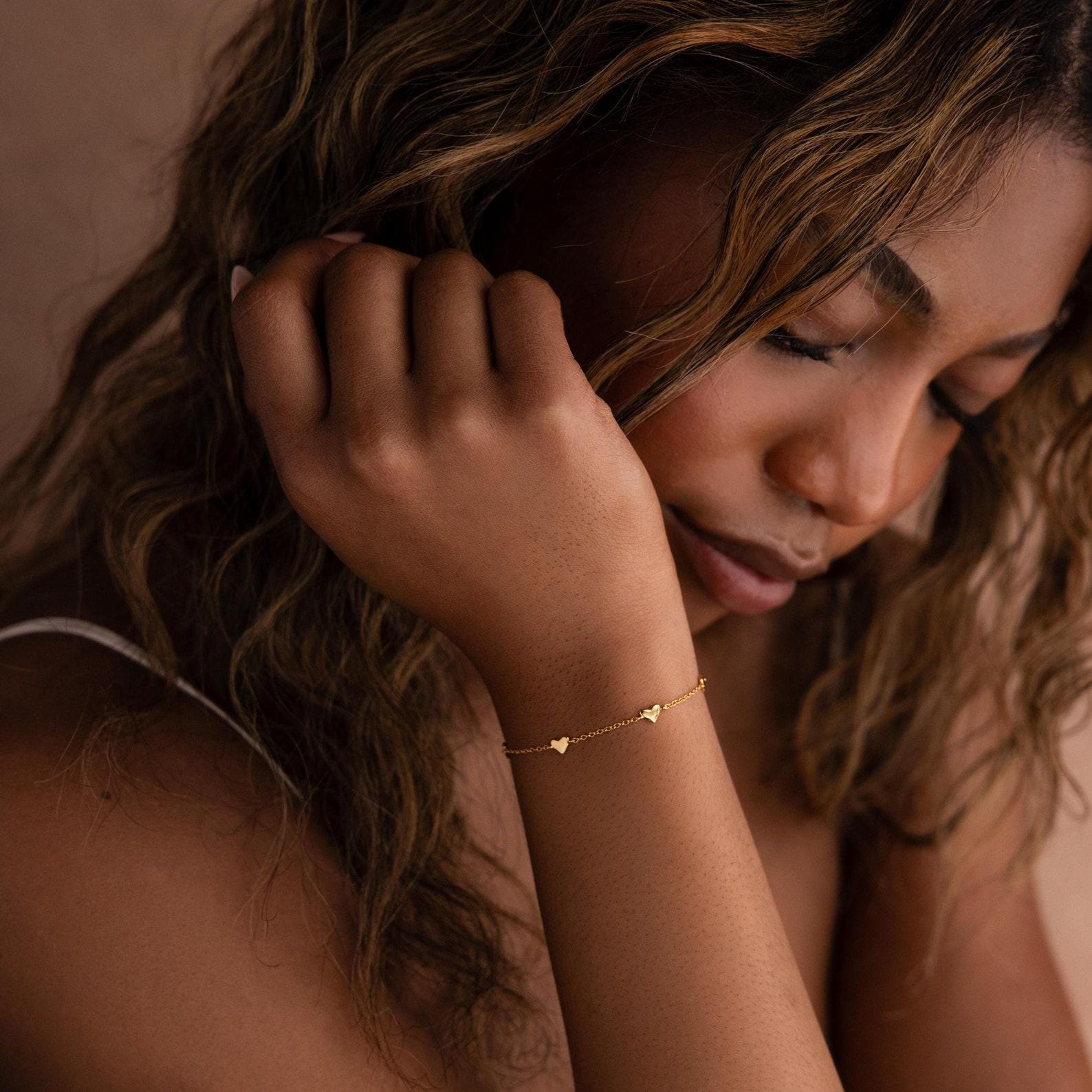Woman with wavy hair wearing the Heart Station Necklace & Bracelet Set in 18K Gold, featuring delicate heart charms, resting her head on her hand.
