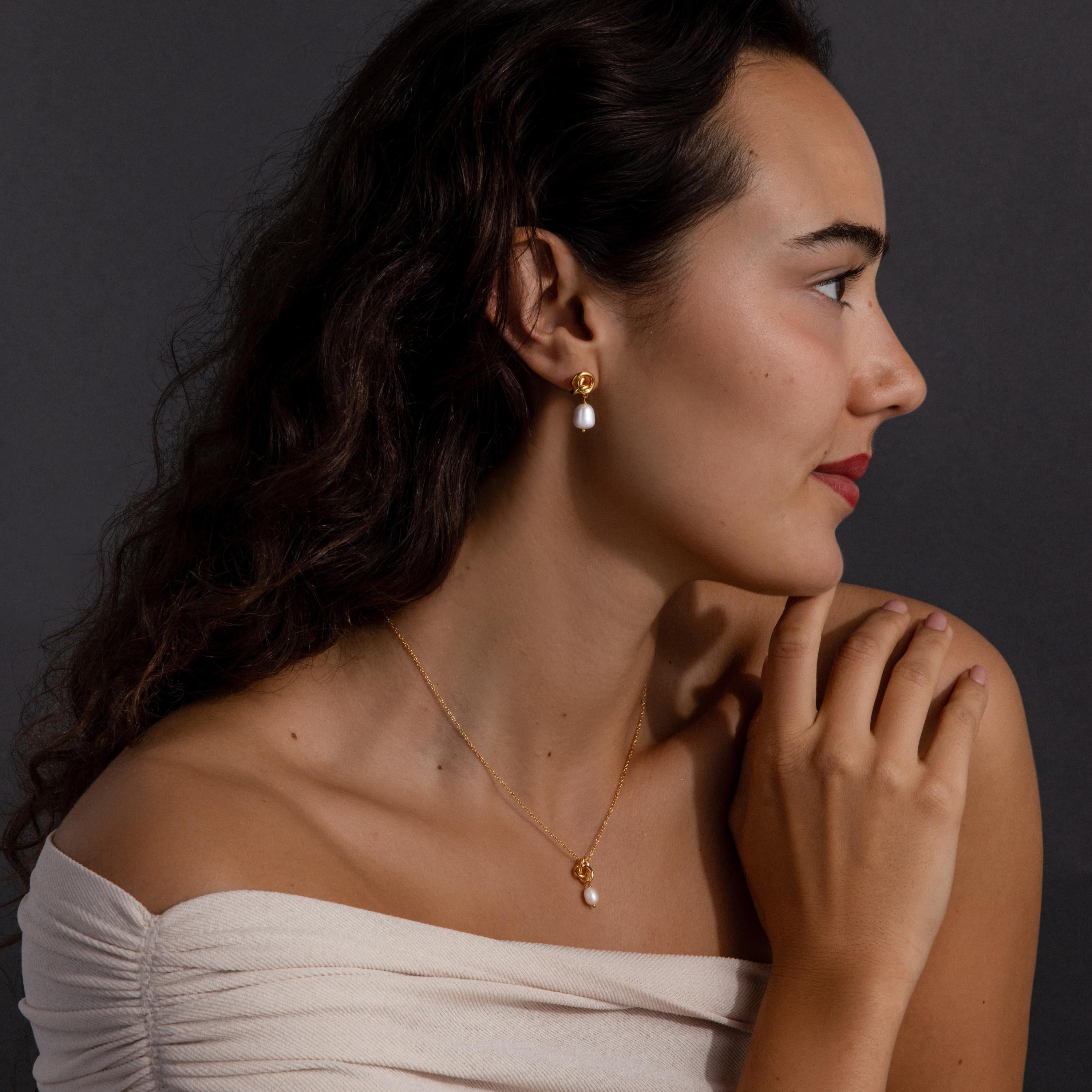 Woman in off-shoulder top wearing the Pearl Knot Necklace & Earrings Set, featuring gold and pearl accents, poses against a dark background—an elegant look, perfect as a gift for your wife.