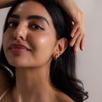 Woman with long dark hair and gold hoop earrings poses with hand resting on her head, exuding confidence in modern Baguette Birthstone Drop Studs.
