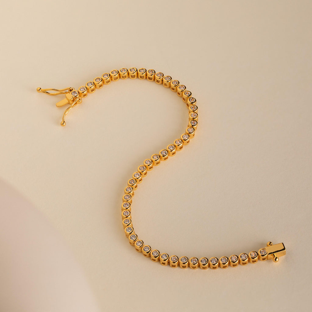 The Diamond Bezel Tennis Necklace & Bracelet Set in 18K Gold is displayed on a beige surface in an elegant S shape, showcasing its round diamonds.