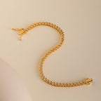 The Diamond Bezel Tennis Necklace & Bracelet Set in 18K Gold is displayed on a beige surface in an elegant S shape, showcasing its round diamonds.