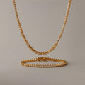 Diamond Bezel Tennis Necklace & Bracelet Set in 18K Gold, featuring round white stones, displayed on a plain beige background.