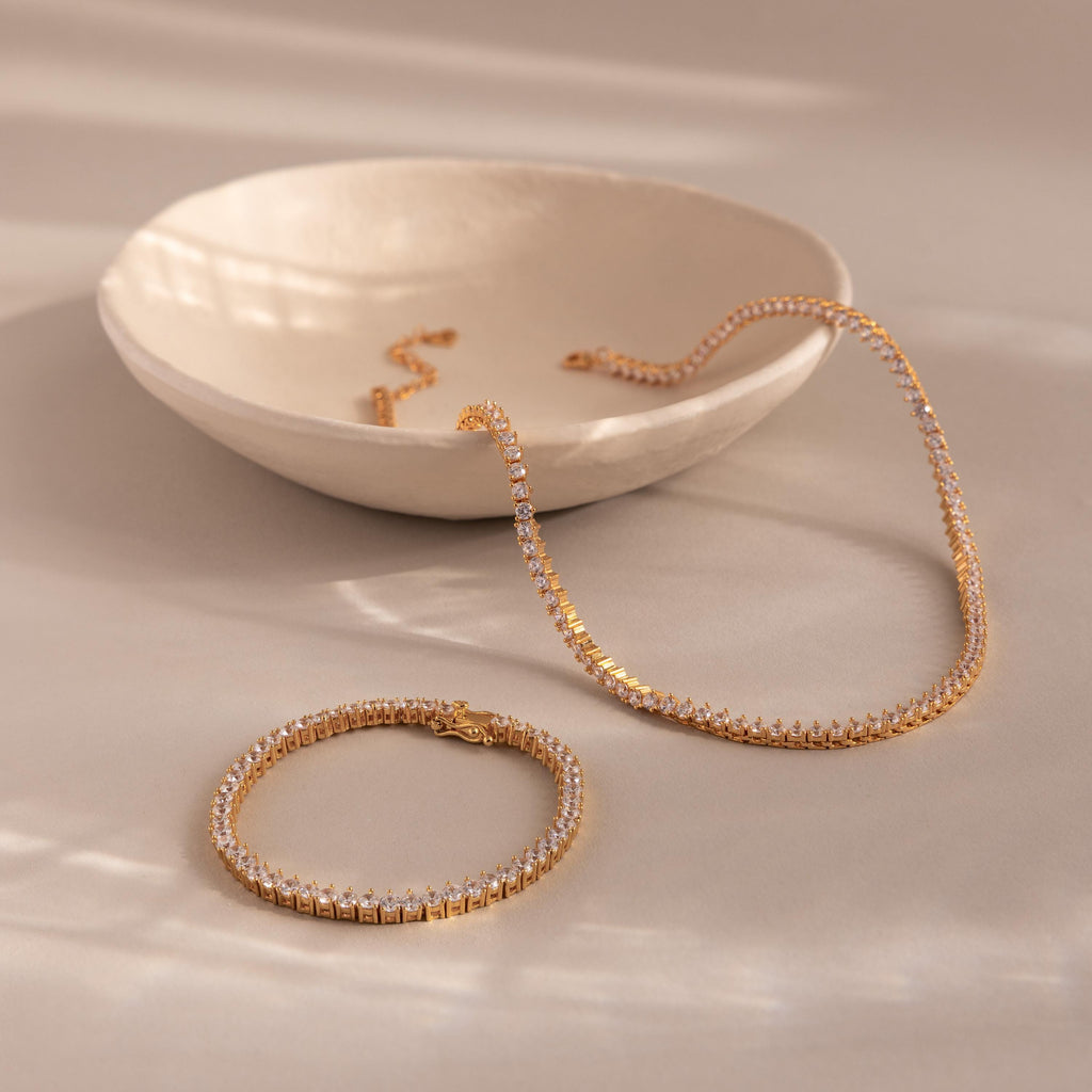 The 3-Prong Diamond Tennis Necklace & Bracelet Set with clear gemstones is displayed on a beige surface next to a ceramic bowl.