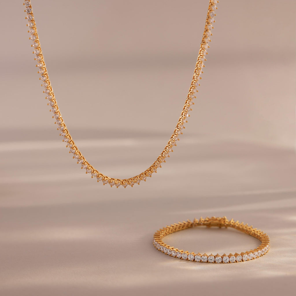 The 3-Prong Diamond Tennis Necklace & Bracelet Set in 18K Gold is displayed on a soft beige surface, with its sparkling diamonds and elegant gold design set against a blurred background.