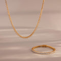 The 3-Prong Diamond Tennis Necklace & Bracelet Set in 18K Gold is displayed on a soft beige surface, with its sparkling diamonds and elegant gold design set against a blurred background.