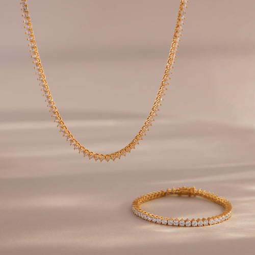 The 3-Prong Diamond Tennis Necklace & Bracelet Set in 18K Gold is displayed on a soft beige surface, with its sparkling diamonds and elegant gold design set against a blurred background.