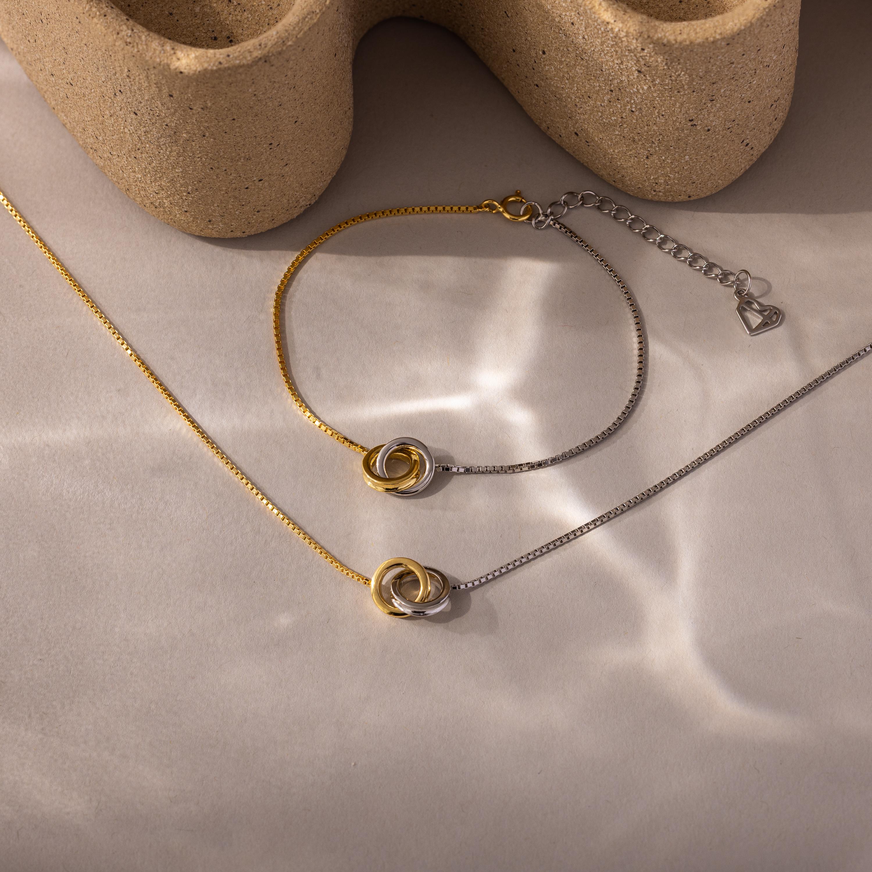 The Interlocking Circles Necklace and Bracelet Set, featuring gold and silver circles, rests on a neutral surface with soft shadows, highlighting this minimalist jewelry set.