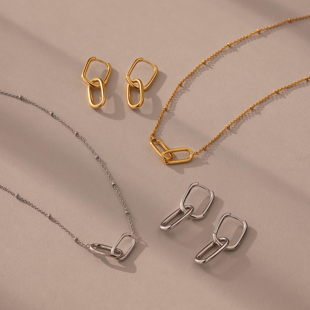The Linked Charm Necklace and Earrings Set is displayed on a beige surface with soft shadows, making it the perfect anniversary gift.