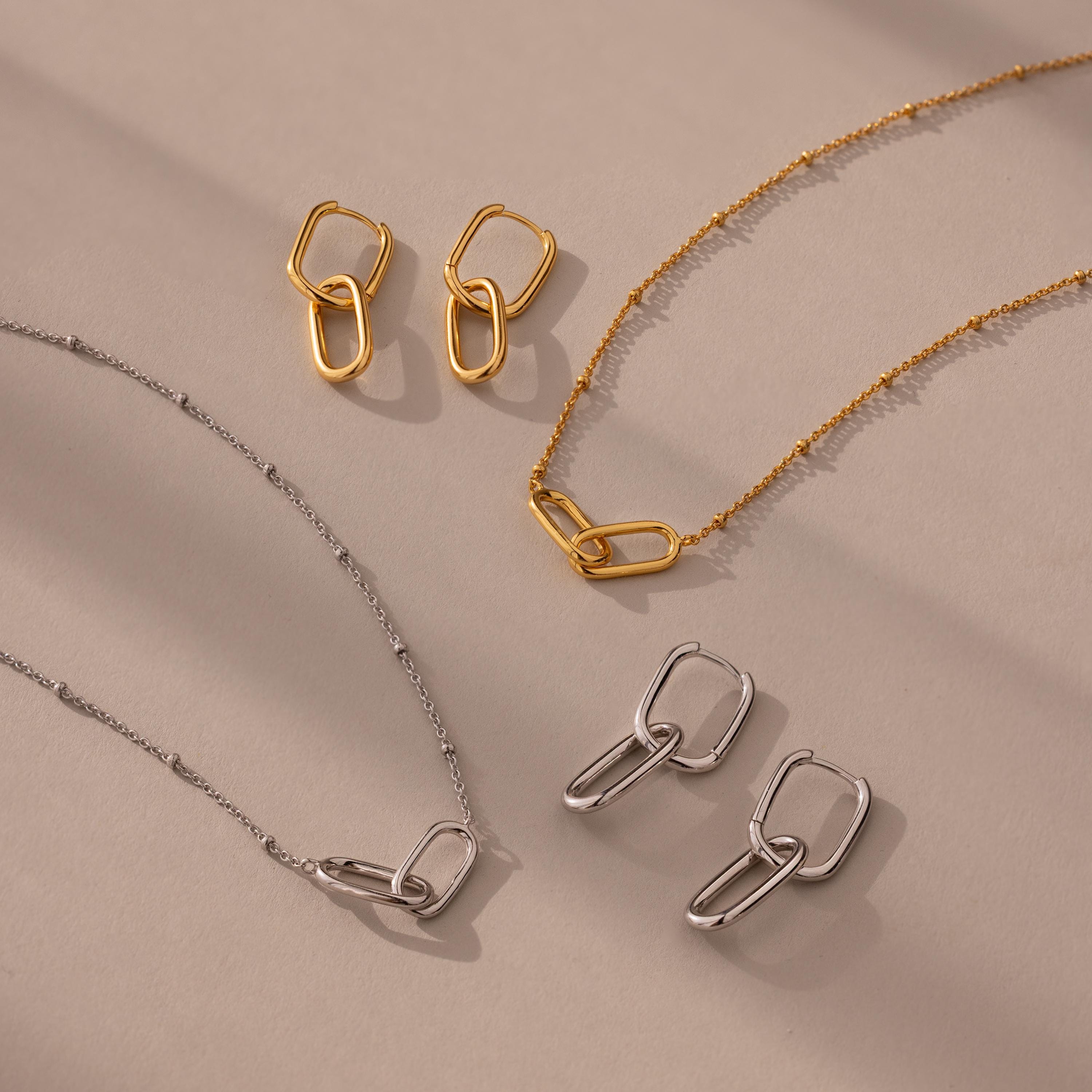 The Linked Charm Necklace and Earrings Set is displayed on a beige surface with soft shadows, making it the perfect anniversary gift.