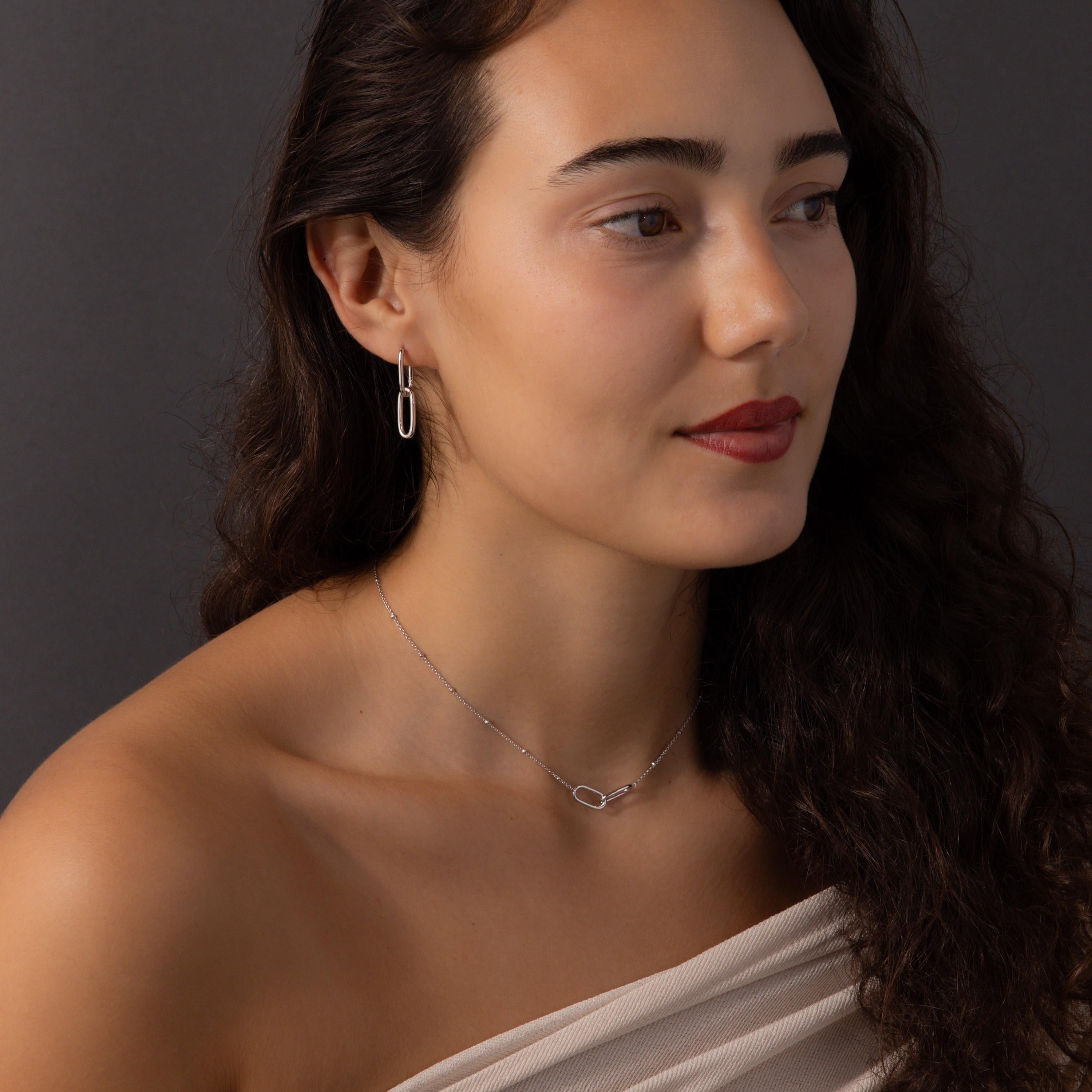 Woman with wavy dark hair wearing the Linked Charm Necklace and Earrings Set in Sterling Silver, gazing to the side against a dark backdrop—an ideal anniversary gift.