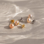 Two pairs of Pink Pearl Studs, one with gold backs and one with silver, are displayed on a textured beige surface in a soft blush tone.