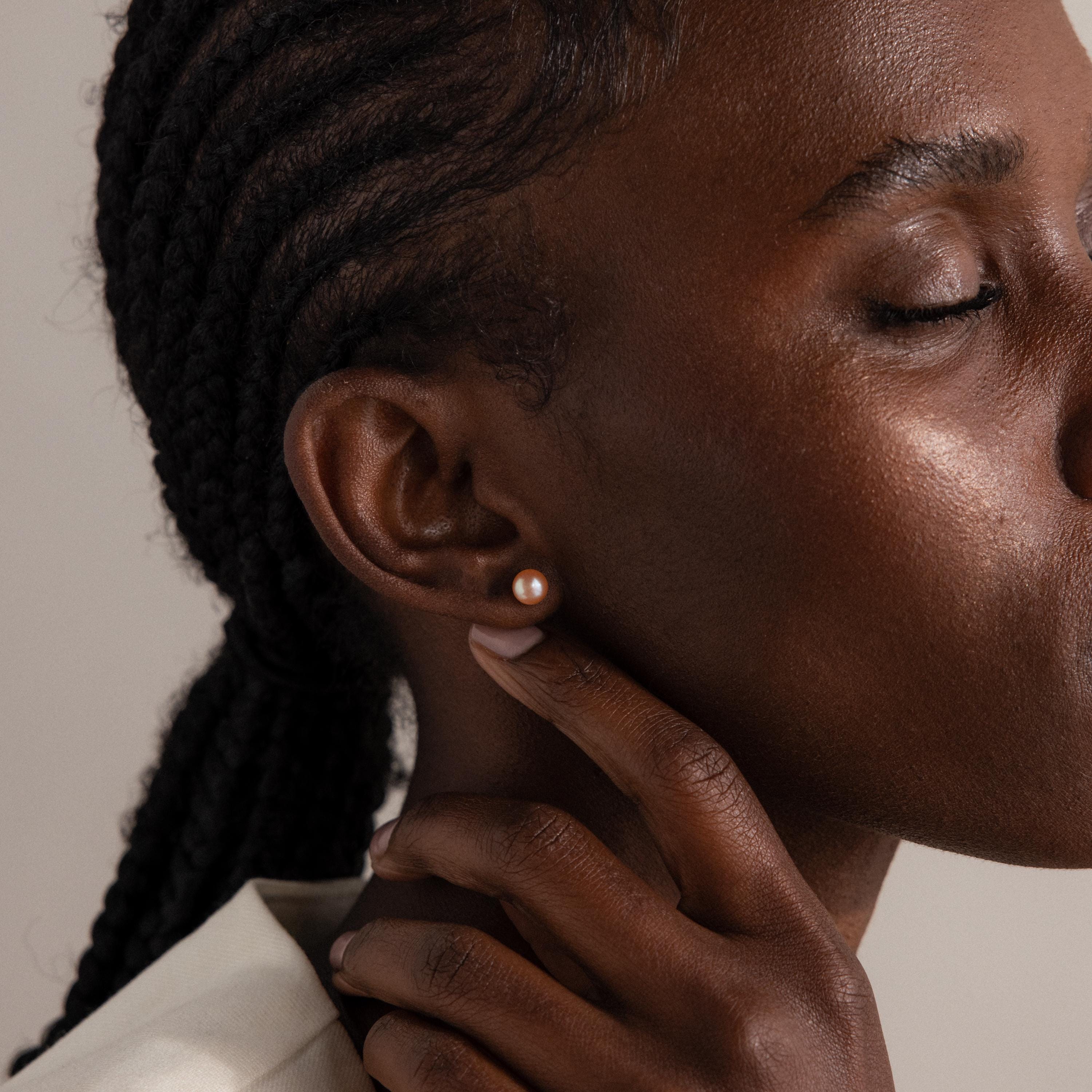 A close-up of a person with braided hair wearing Pink Pearl Studs in 18K Gold, eyes closed and hand gently touching their neck.