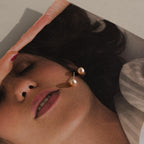 A pair of Pink Pearl Studs in a blush tone rests on a magazine photo featuring a woman's face with closed eyes.