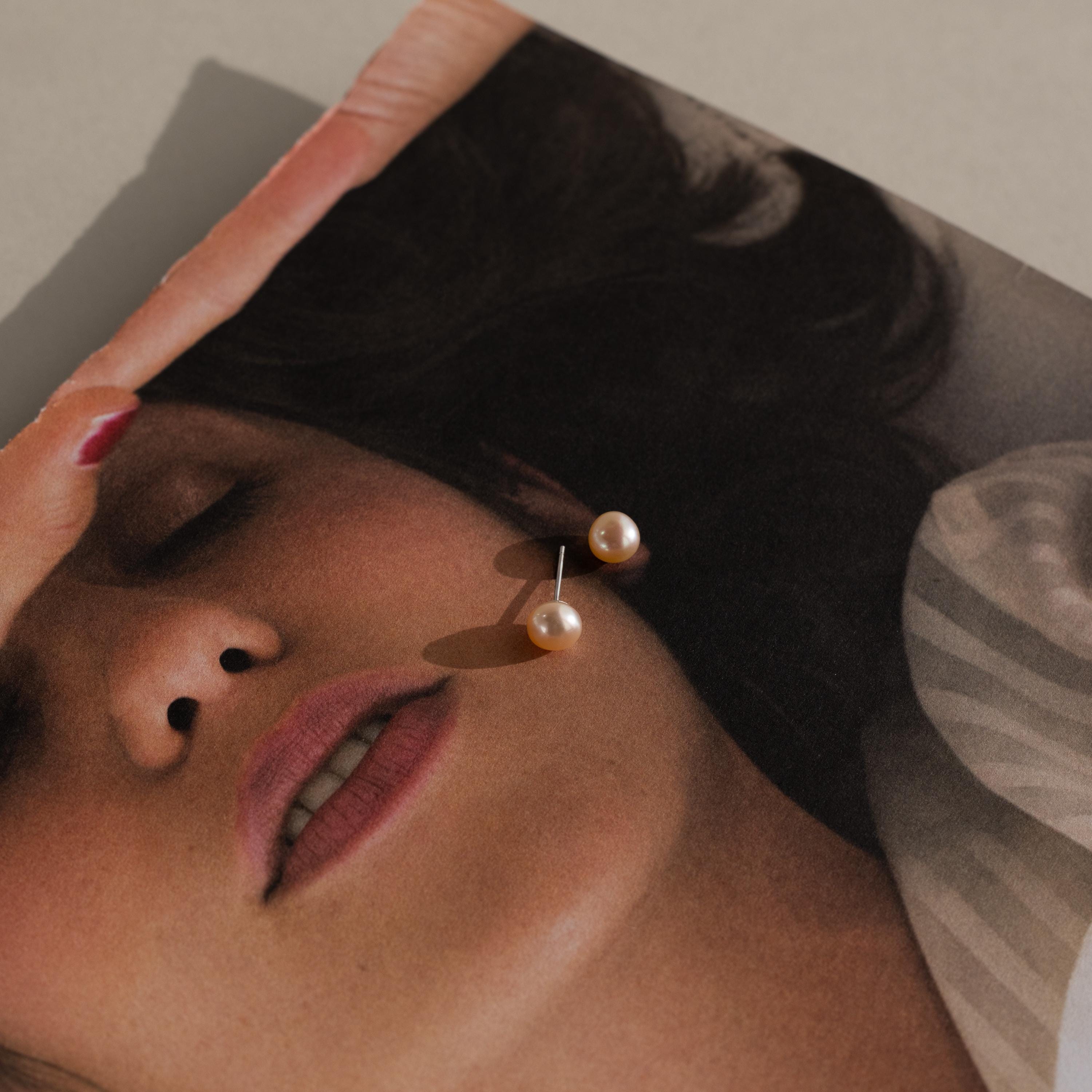 A pair of Pink Pearl Studs in a blush tone rests on a magazine photo featuring a woman's face with closed eyes.
