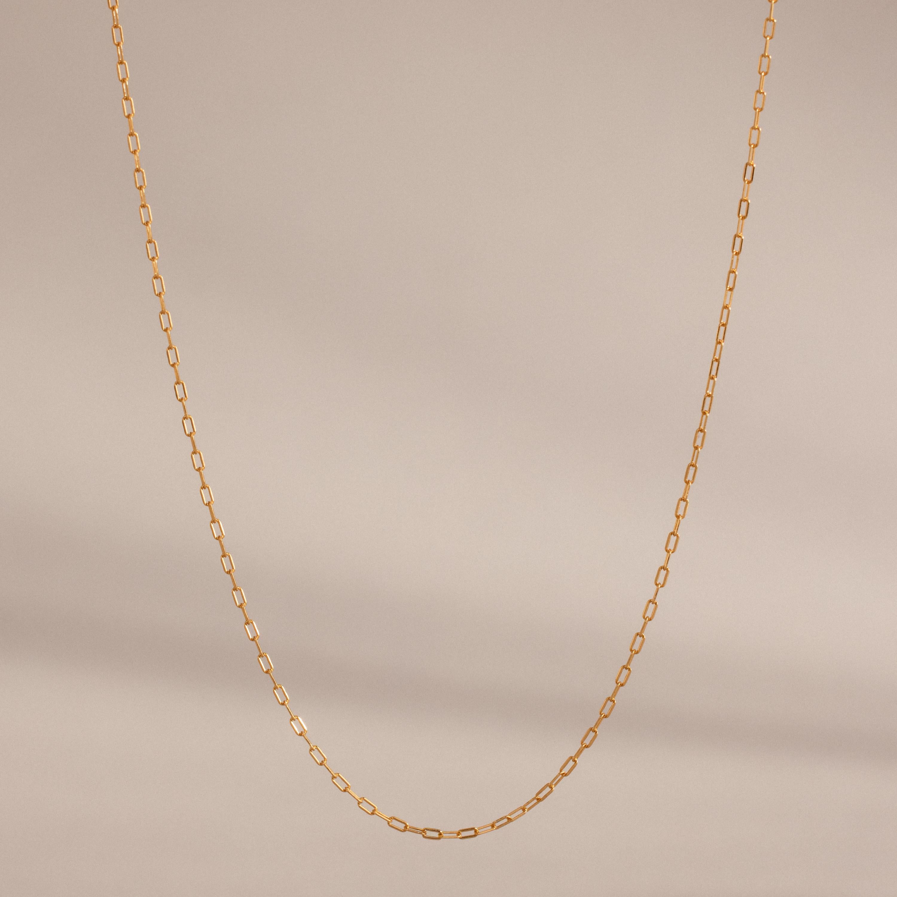 The Dainty Paperclip Necklace features small rectangular gold links, ideal for layering as a simple, elegant accessory, shown against a plain beige background.