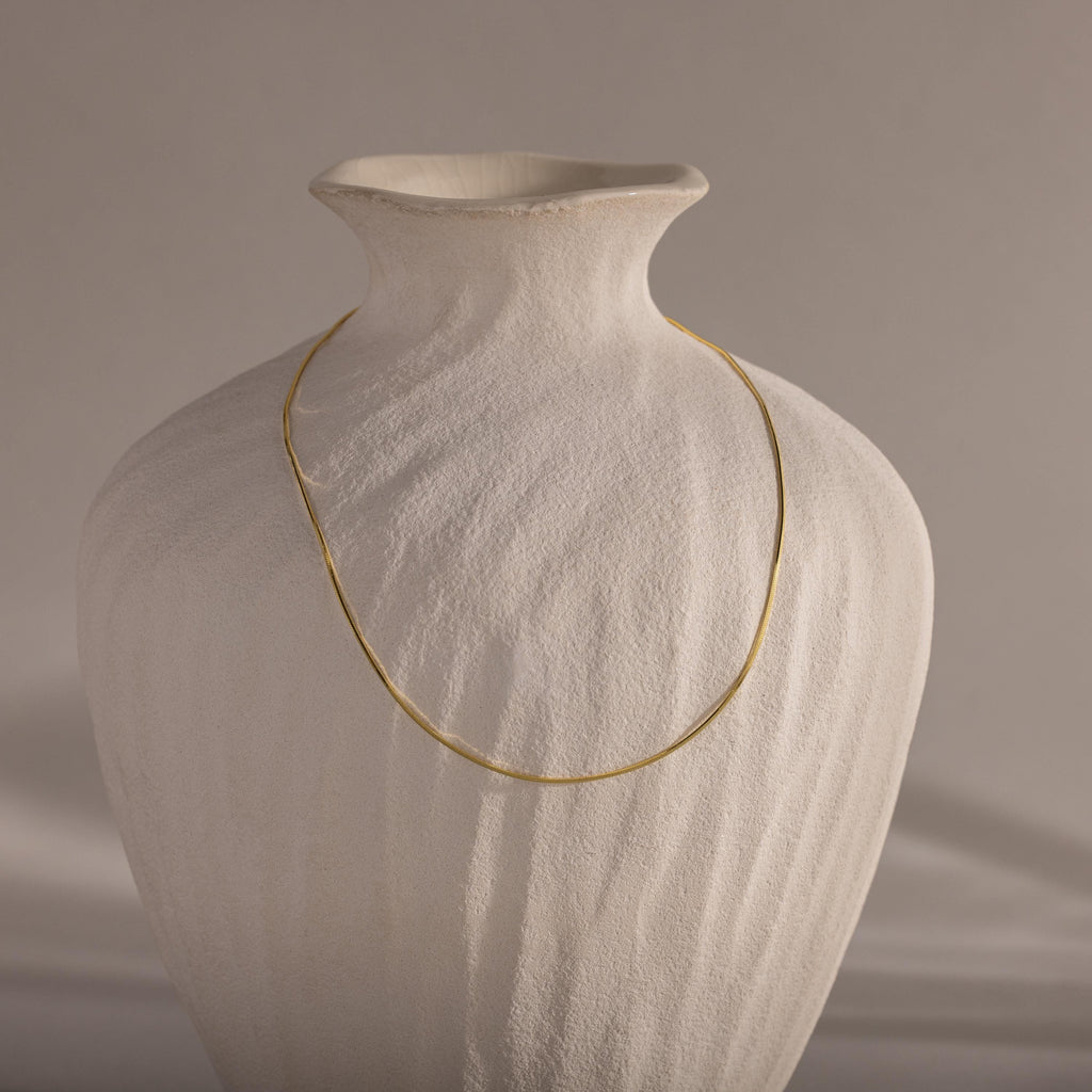 The Dainty Herringbone Necklace in 18K Gold is elegantly draped over a textured white ceramic vase, set against a neutral background.