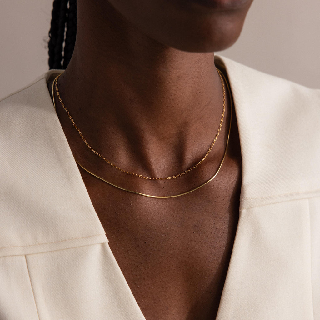 A person wearing the Dainty Herringbone Necklace in 18K Gold layered with another gold necklace, paired with a cream-colored v-neck top.
