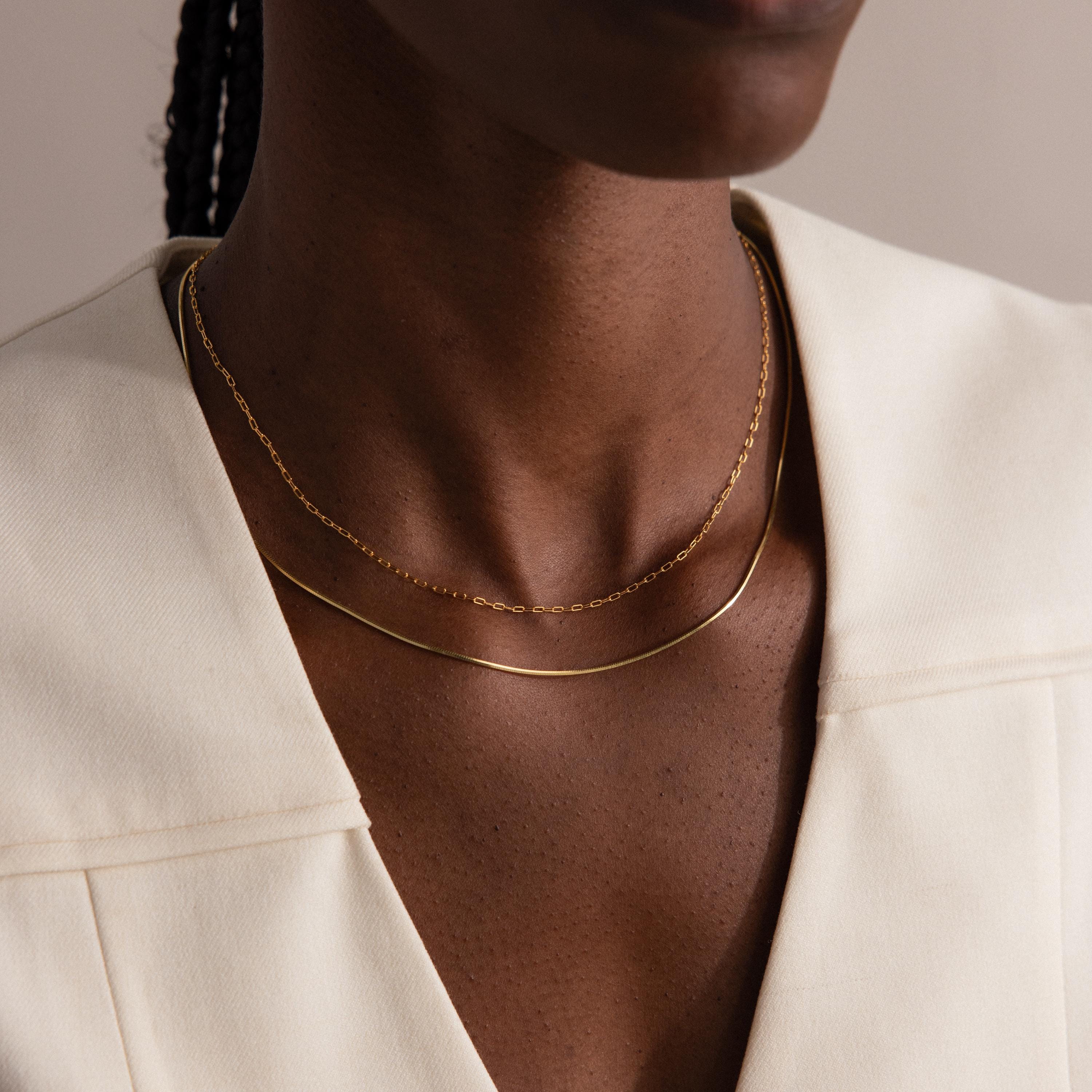 A person wearing the Dainty Herringbone Necklace in 18K Gold layered with another gold necklace, paired with a cream-colored v-neck top.