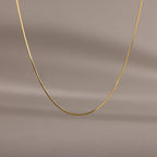 The Dainty Herringbone Necklace features a delicate curve, beautifully displayed against a simple beige background.