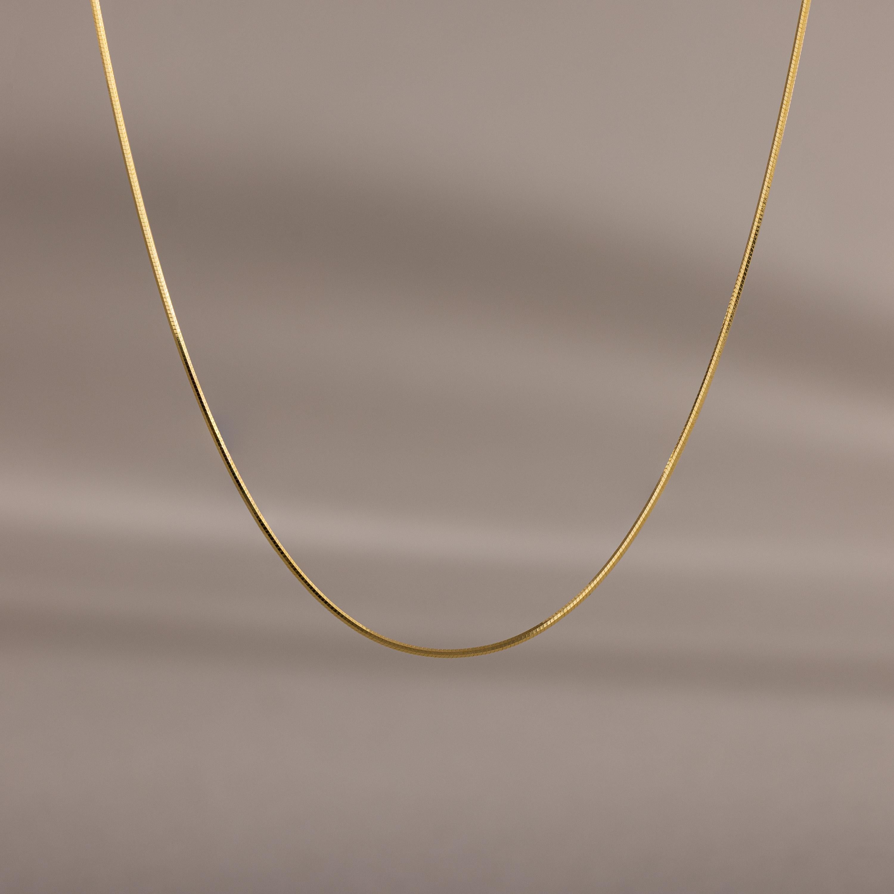 The Dainty Herringbone Necklace features a delicate curve, beautifully displayed against a simple beige background.