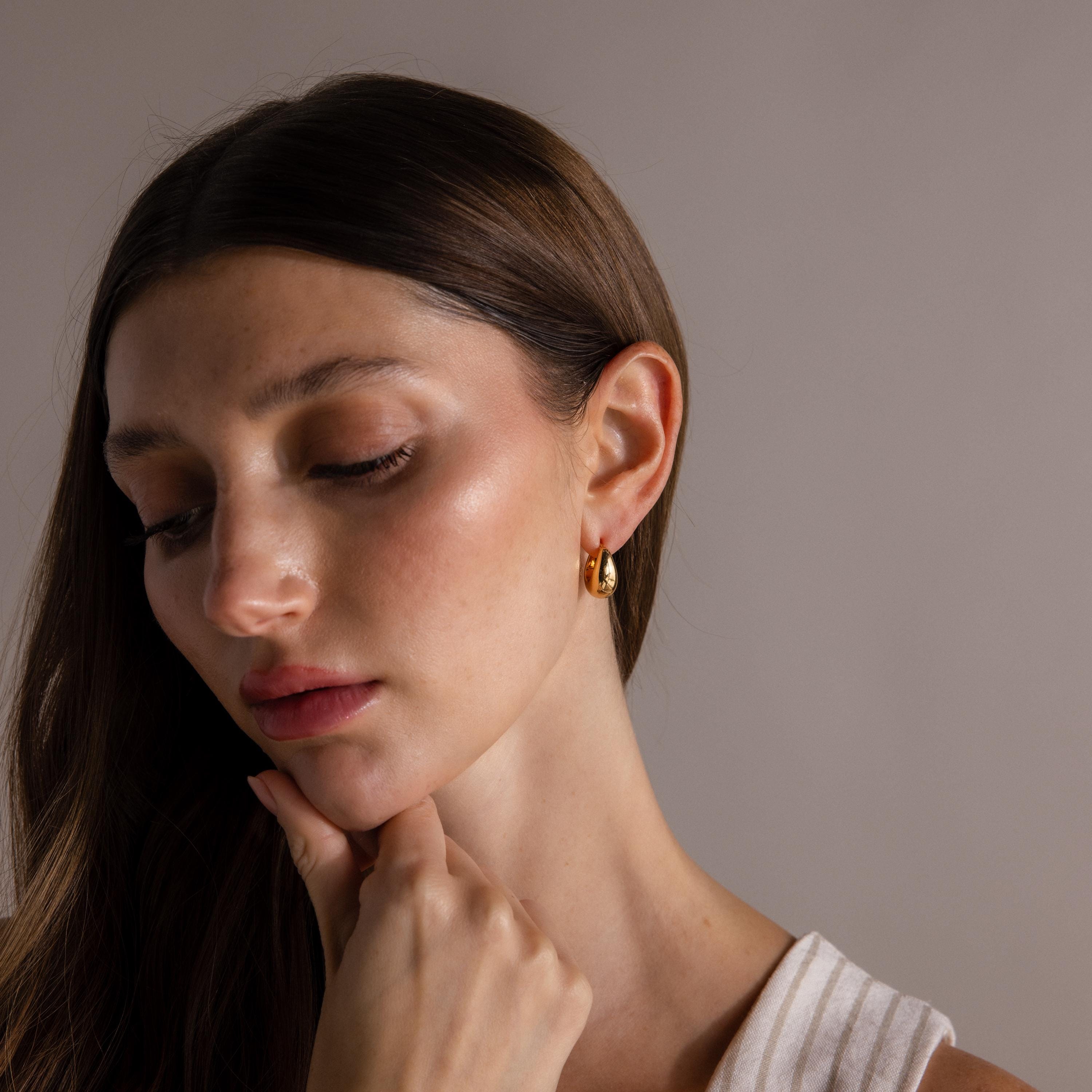 A woman with long brown hair looks down, her hand touching her chin, wearing Bold Teardrop Earrings in 18K Gold with a high polish finish.