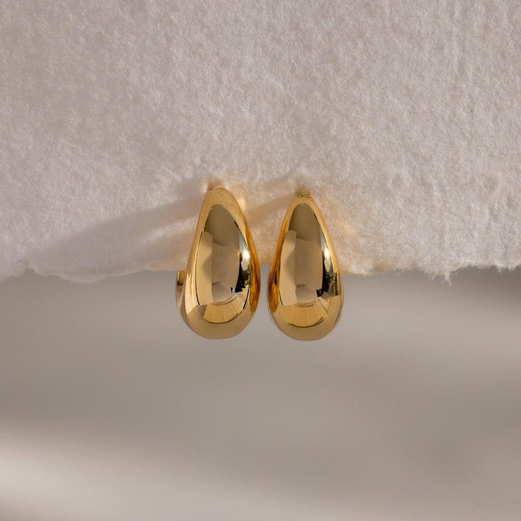 The Bold Teardrop Earrings, featuring a shiny gold finish, rest on a white textured surface—ideal for adding a modern touch to any outfit.