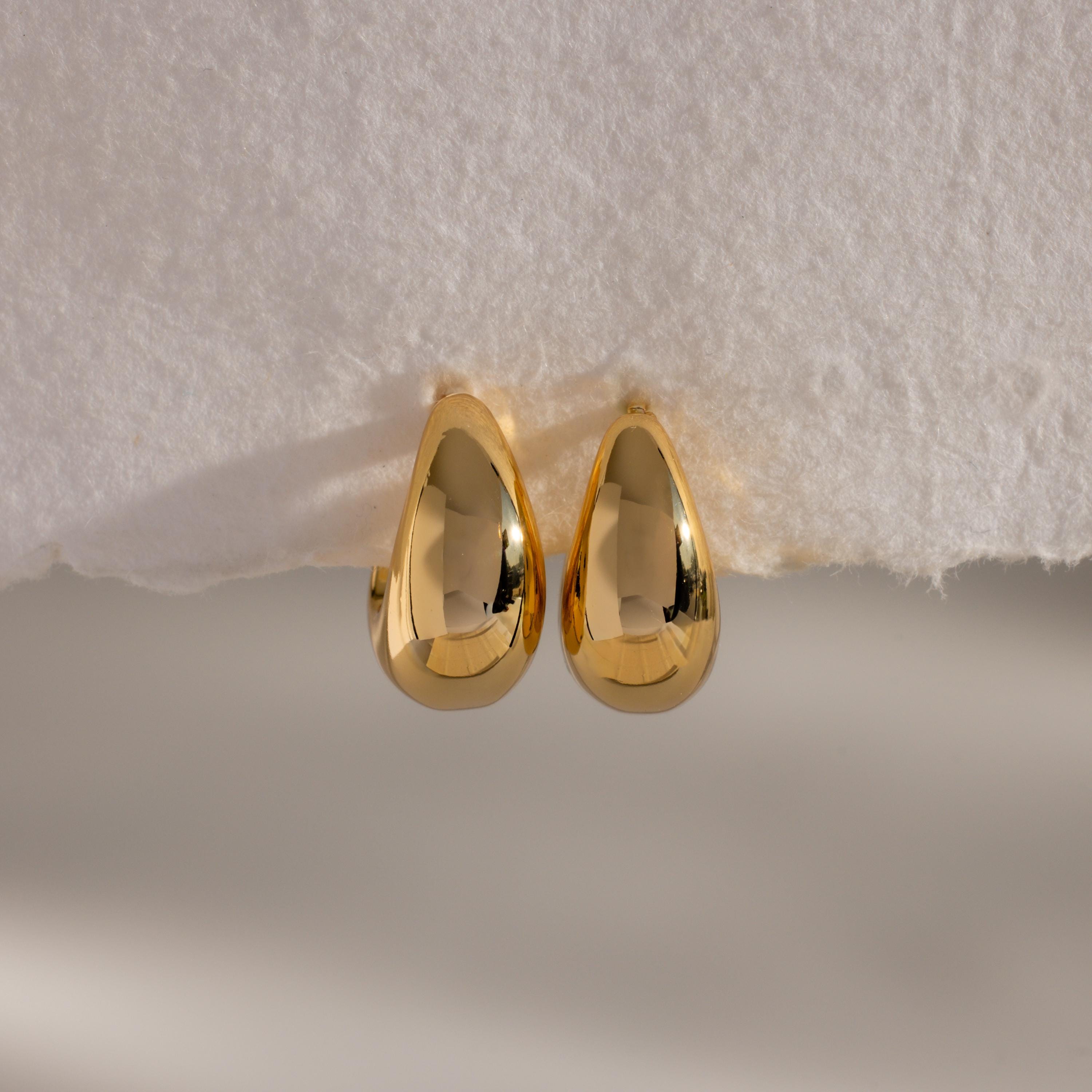 The Bold Teardrop Earrings, featuring a shiny gold finish, rest on a white textured surface—ideal for adding a modern touch to any outfit.