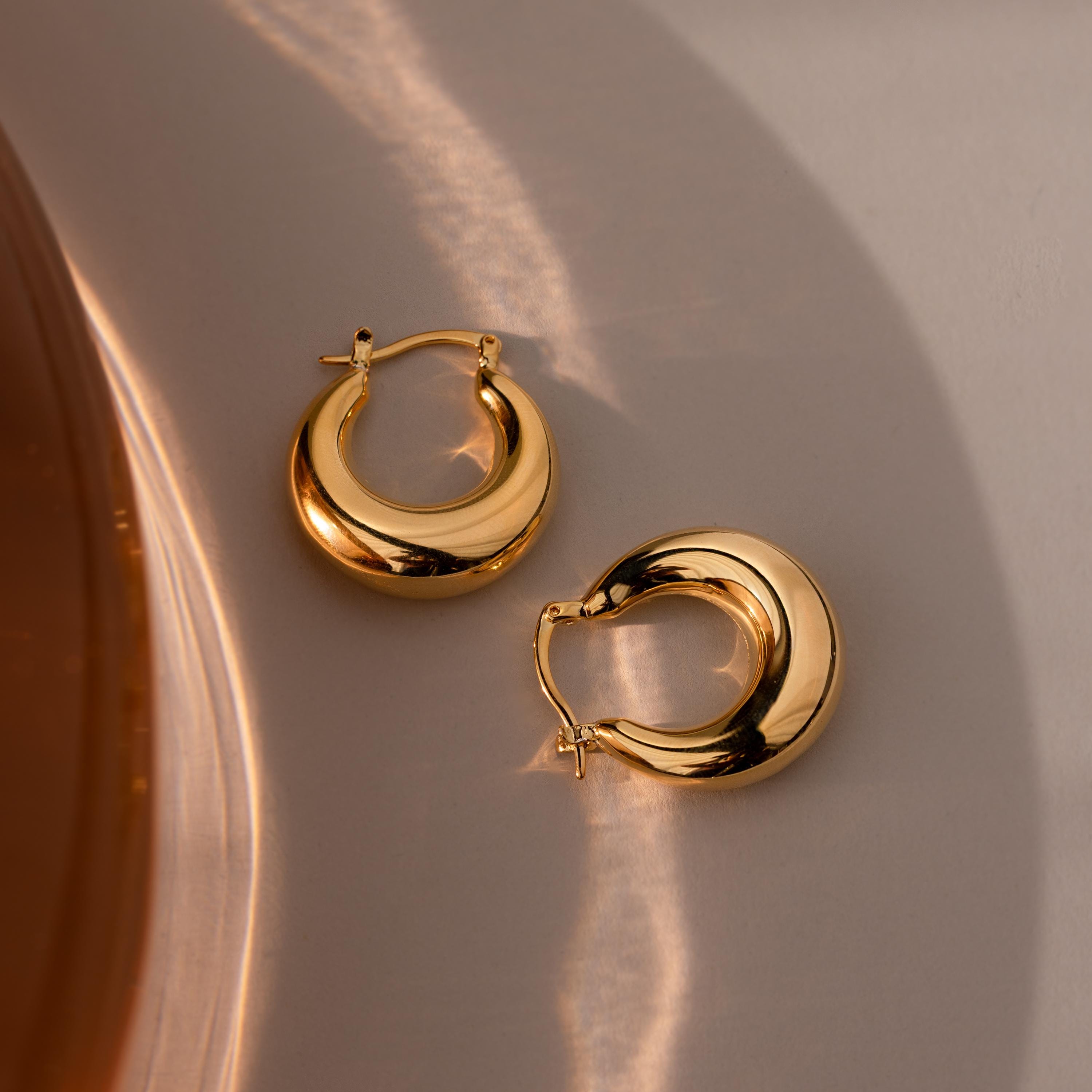 Sculpted Hoops in 18K Gold rest on a neutral surface with gentle light and shadow—modern classic earrings that reflect timeless elegance.