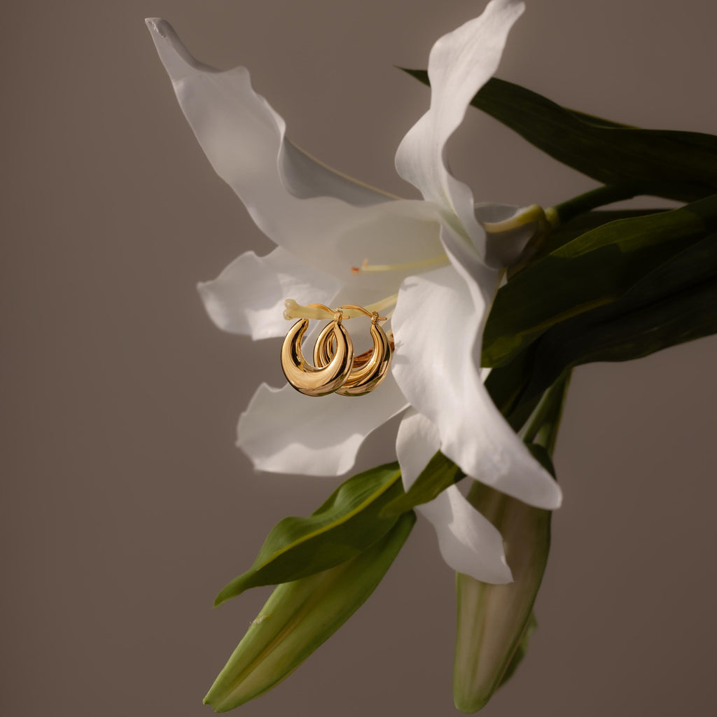 Sculpted Hoops in 18K Gold hang on a blooming white lily with green leaves, radiating timeless elegance against a soft background.