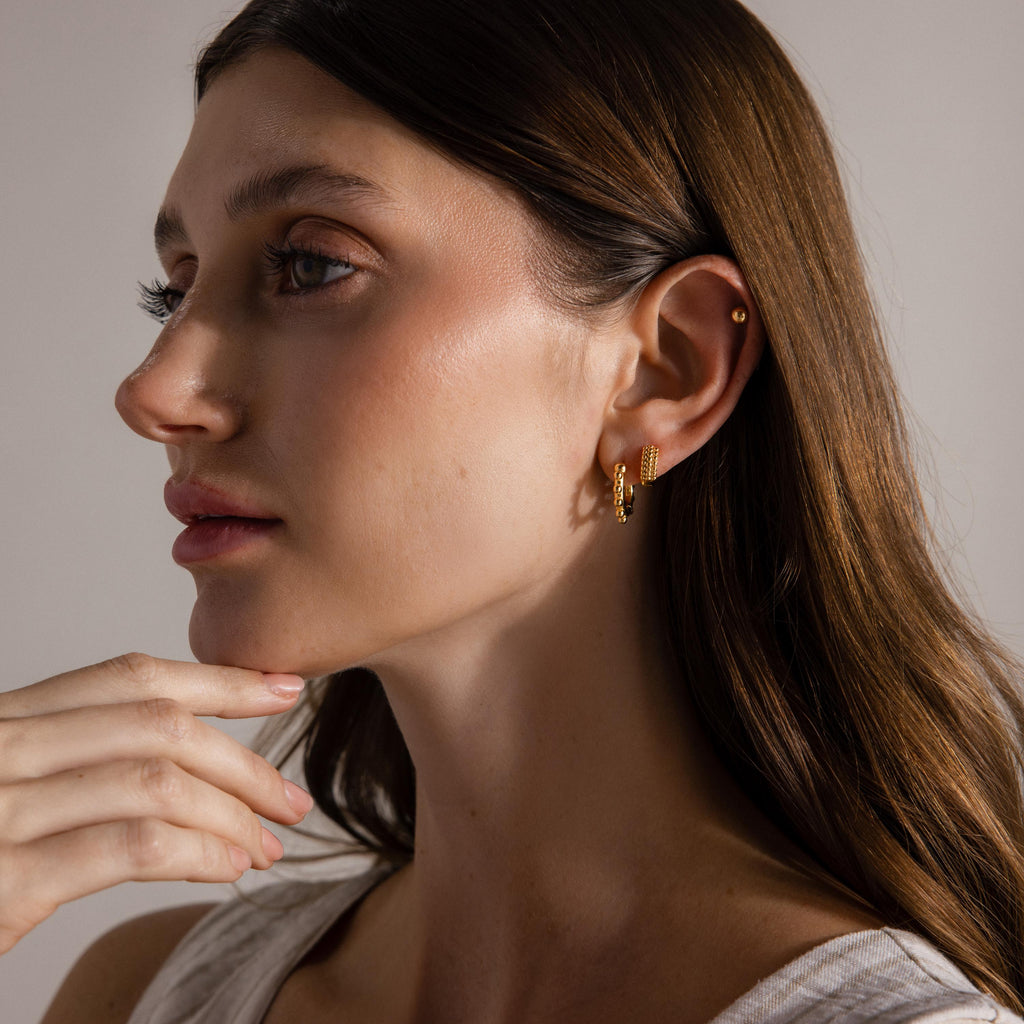 Woman with long brown hair wearing Beaded Chunky Hoops in 18K Gold, gazing to the side with her hand near her chin.