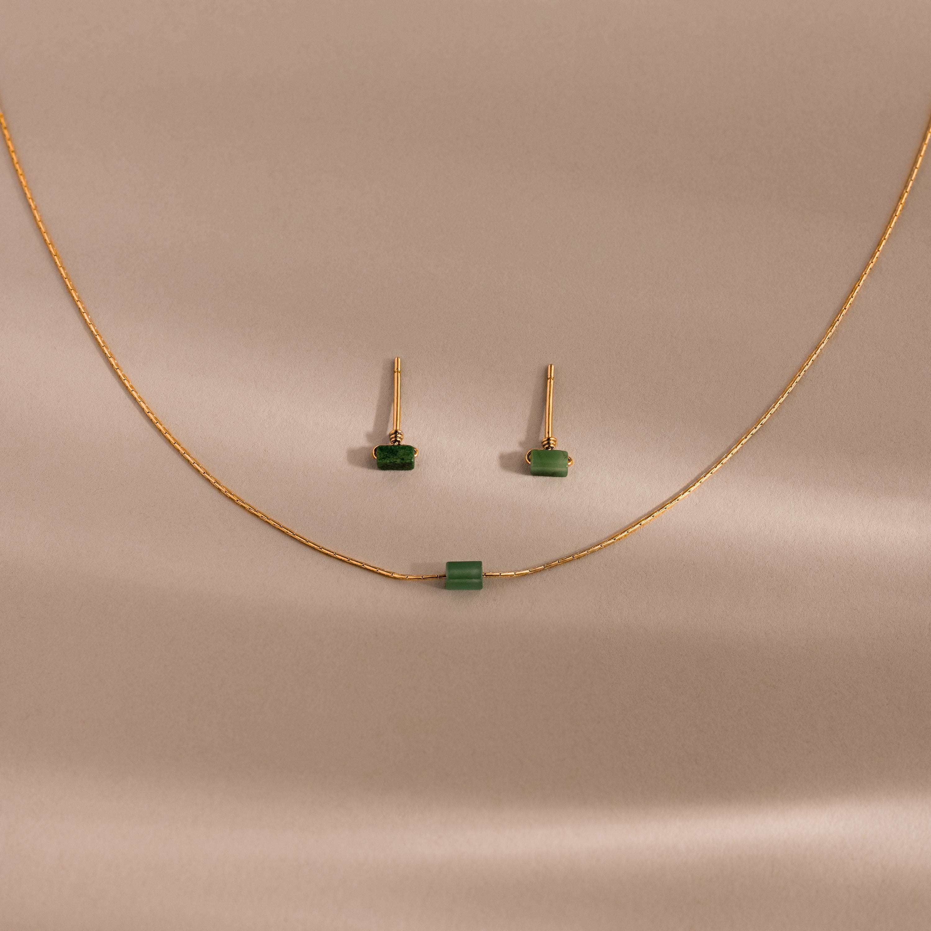 The Jade Bar Jewelry Set features a gold necklace and earrings with petite green jade stones, displayed on a beige background—an elegant accessory to enhance any outfit.
