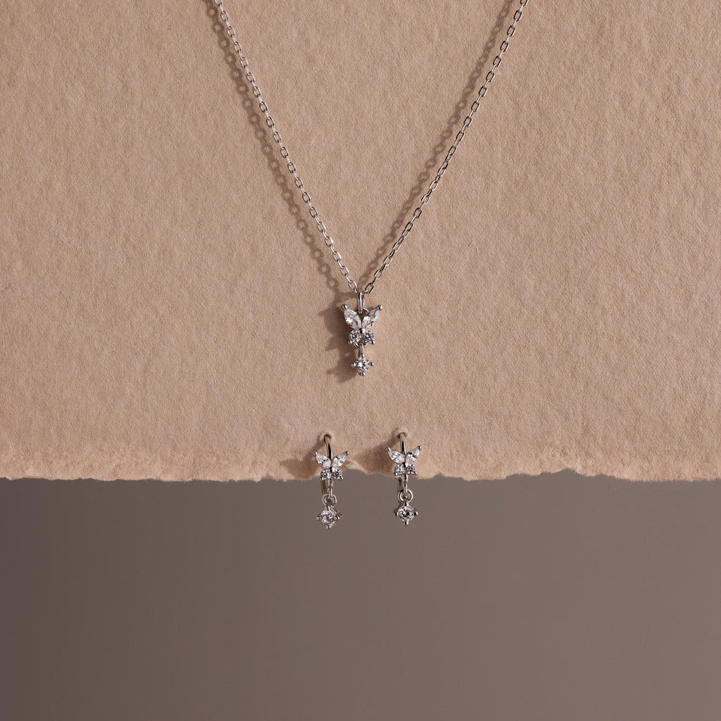 Diamond Butterfly Drop Jewelry Set in Sterling Silver, featuring a necklace and matching huggie earrings with butterfly-shaped pendants, displayed on textured beige paper.