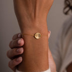 The Dainty Organic Fingerprint Bracelet, featuring a round disc charm, rests elegantly on a person’s wrist as their hands gently touch this unique keepsake jewelry piece.