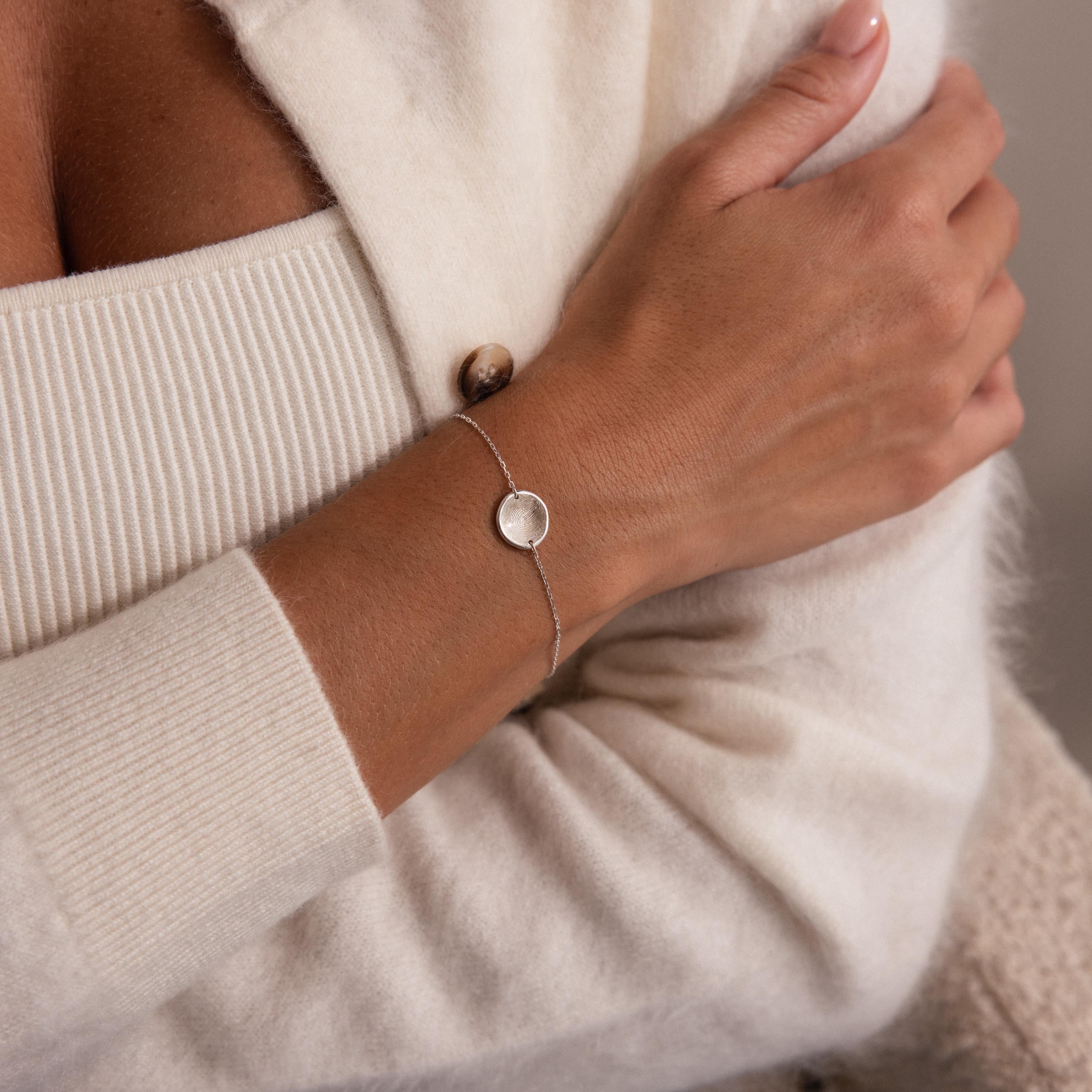 A woman wearing the Dainty Organic Fingerprint Bracelet with a round pendant, paired with a soft cream sweater—a timeless keepsake jewelry piece.
