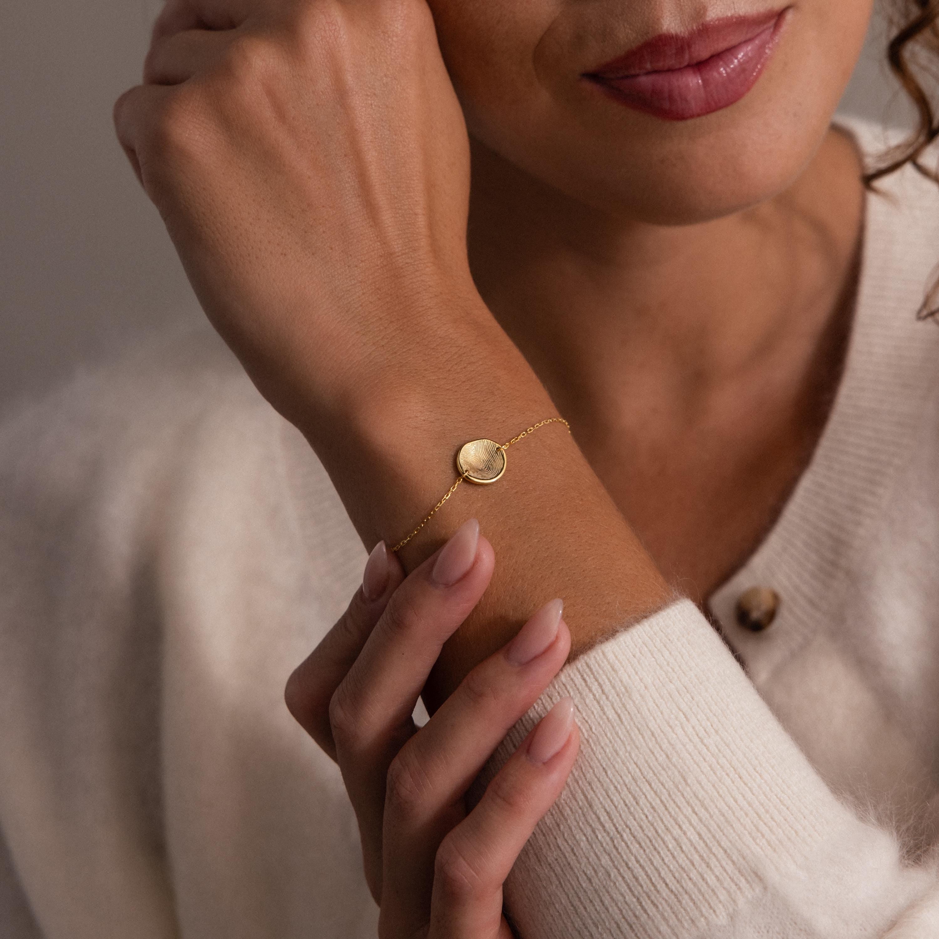 A woman in a cream sweater gently touches her wrist, showcasing the Dainty Organic Fingerprint Bracelet—a beautiful piece of keepsake jewelry.