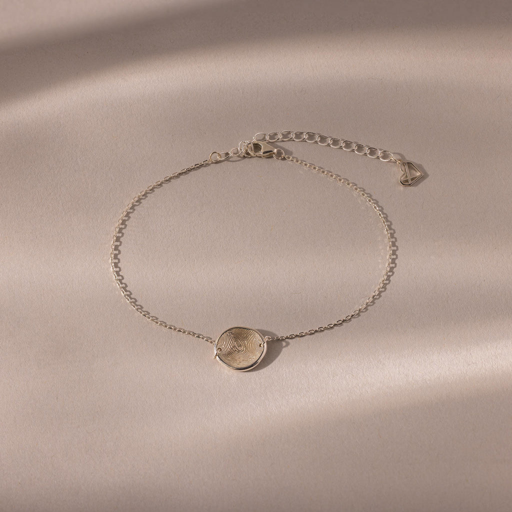 The Dainty Organic Fingerprint Bracelet in Sterling Silver features a round engraved charm, displayed on a beige textured background with soft shadows—an elegant, personalized jewelry piece.